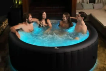 Four people enjoying a relaxing evening in a glowing blue MSPA Aurora inflatable hot tub indoors.