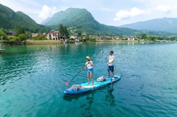 A couple stand-up paddles together on the Aqua Marina SUPER TRIP TANDEM 14’0″ inflatable board, gliding across crystal-clear water with alpine mountains and lakeside villages in the background.