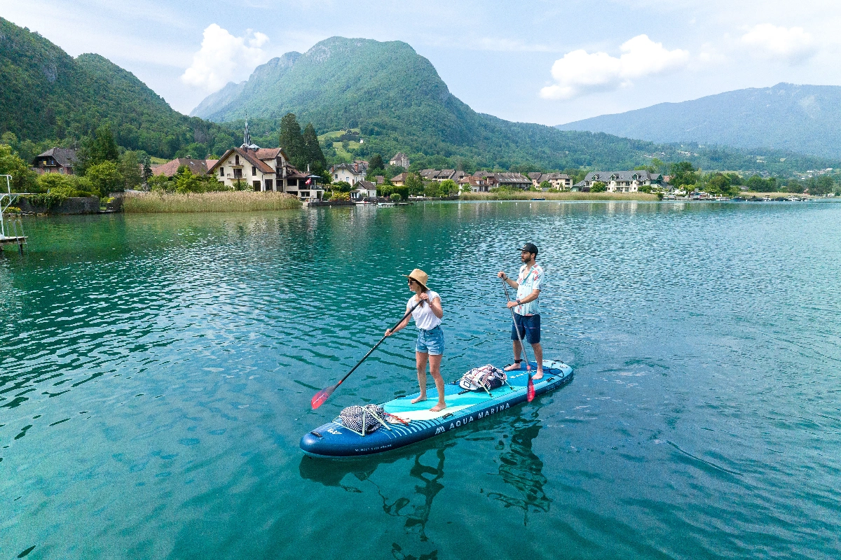 A couple stand-up paddles together on the Aqua Marina SUPER TRIP TANDEM 14’0″ inflatable board, gliding across crystal-clear water with alpine mountains and lakeside villages in the background.