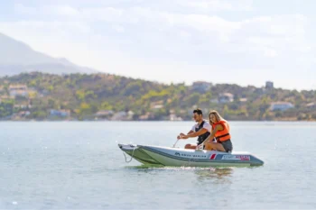 A smiling couple wearing life jackets paddles the Aqua Marina AIRCAT 11’0″ inflatable catamaran across calm, sunlit water with a scenic mountainous shoreline in the background, the sleek white-and-green twin-hull design gliding smoothly as they enjoy a relaxed day on the lake.1.9sFast